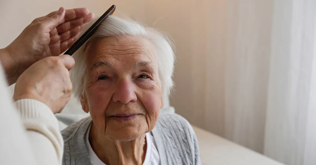 A senior woman sitting in a chair and looking up at another person helping her comb her white hair with a black comb.