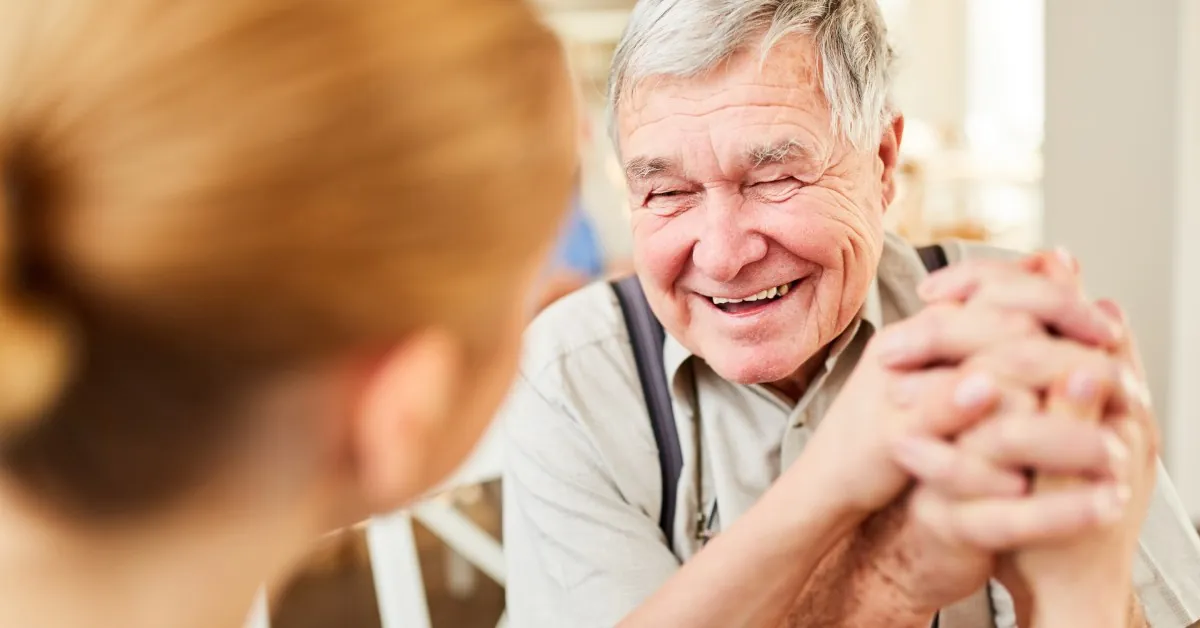 A young woman with brunette hair holding the hands of a senior man with gray hair who is smiling at her.