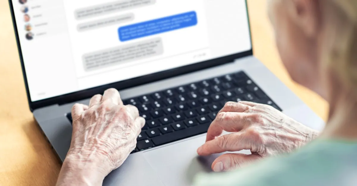 Wrinkled hands type on a silver laptop with blue and grey chat messages visible on the blurred screen.