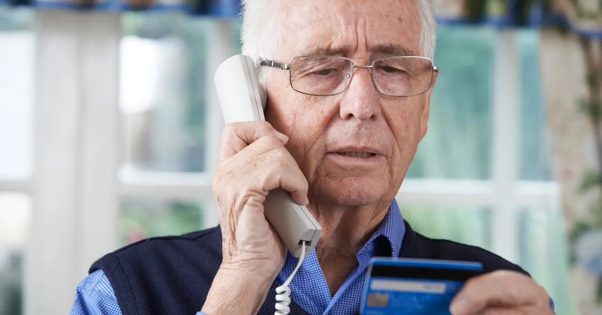A man with white hair holds a landline and blue credit card while looking at the card near curtained windows.