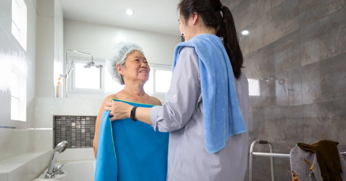 A younger woman holds up a blue towel in front of an older woman standing in front of a white bathtub.