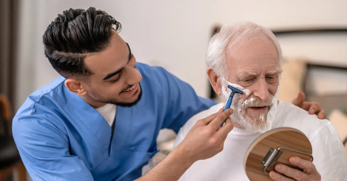 An older man holds a mirror while a younger man in blue scrubs shaves his face with a navy razor and shaving cream.