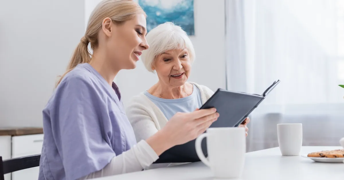 A nurse in purple scrubs sits beside a senior woman at a dining table as they look through a photo book together.