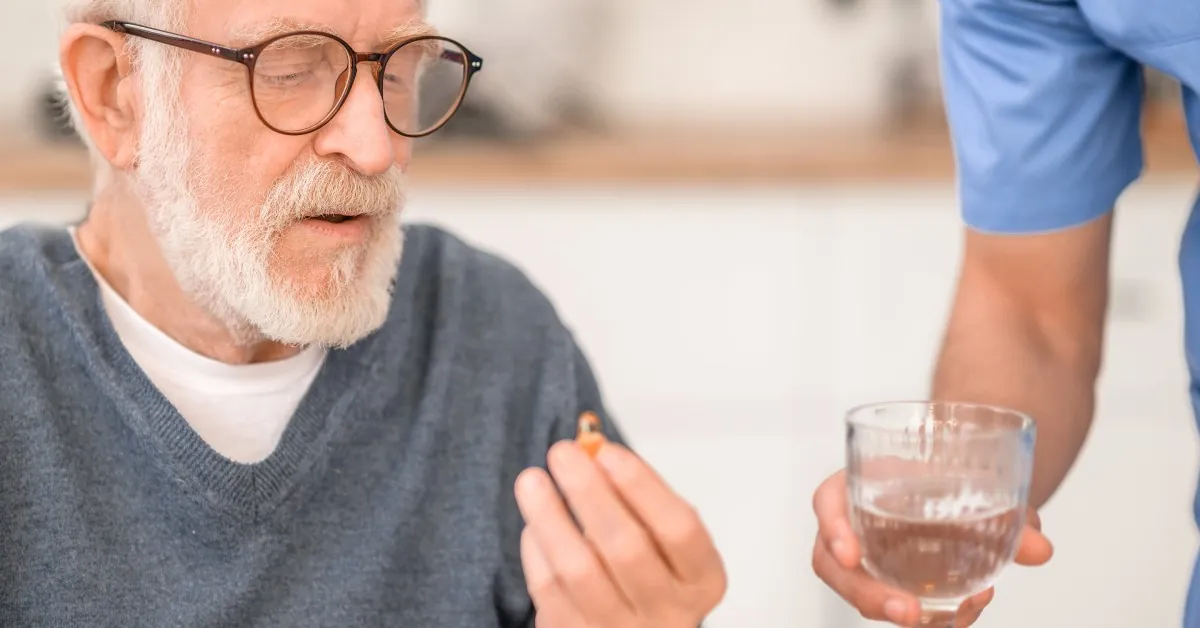 A senior man sits indoors taking medication as a male caregiver hands him a glass of water during routine care.