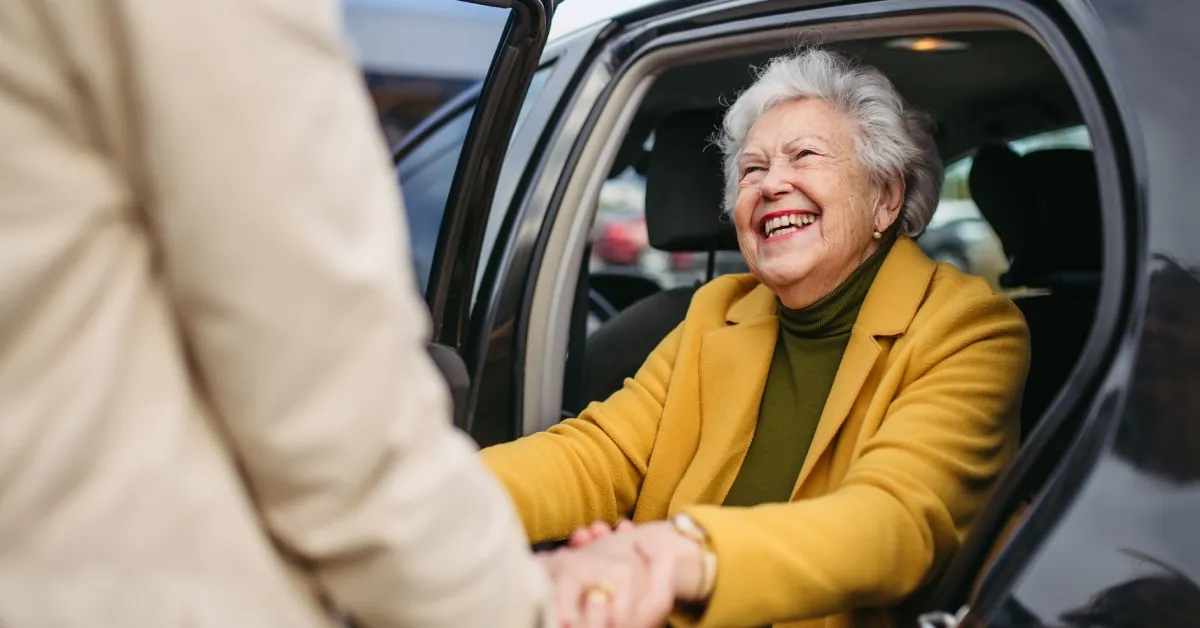 A senior woman smiles as a caregiver helps her stand by the backseat of a car, holding her hands for support.