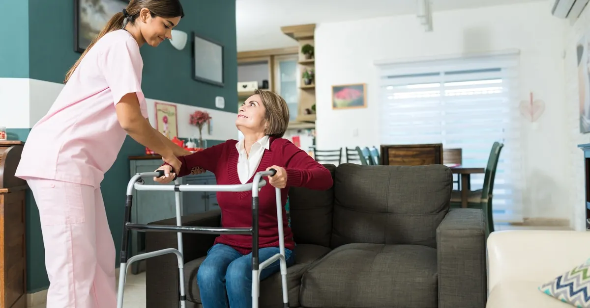 An in-home nurse helps a senior woman stand from a couch using a walker as the woman looks up and smiles.