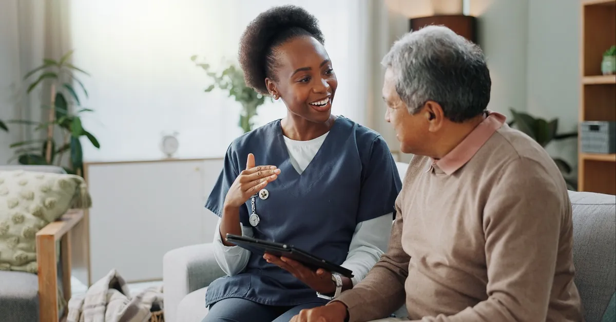 An in-home nurse sits beside a senior man on a couch, holding a tablet as she explains something to him.