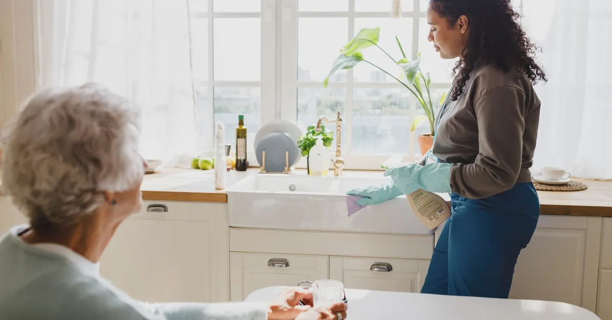 A caregiver wearing gloves cleans a kitchen counter while an elderly woman sits nearby holding a glass of water.