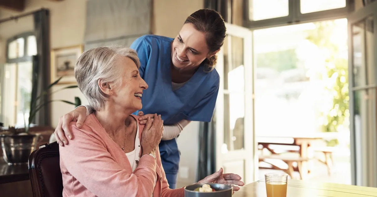 A caregiver in blue scrubs smiles and rests a hand on an elderly woman’s shoulder as she eats breakfast at a table.