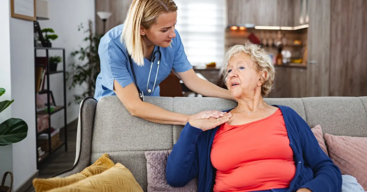 A caregiver checks on an elderly woman resting on a couch, gently assessing her condition in a home care setting.