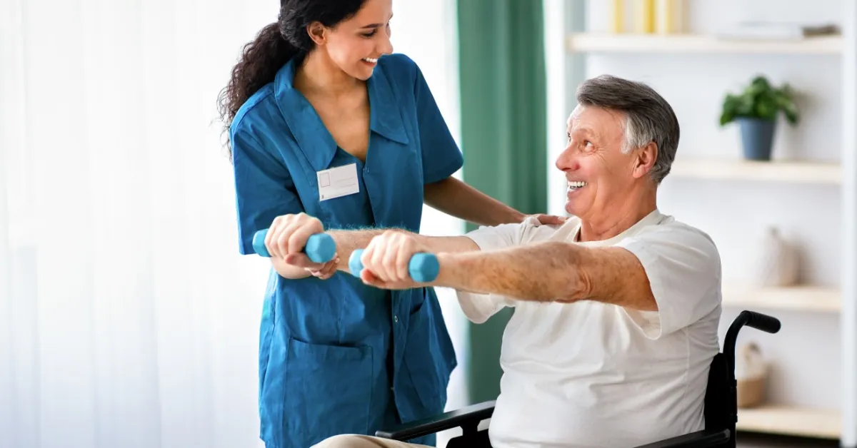 A caregiver assists a senior man in a wheelchair with light-weight exercises, supporting strength and rehabilitation.