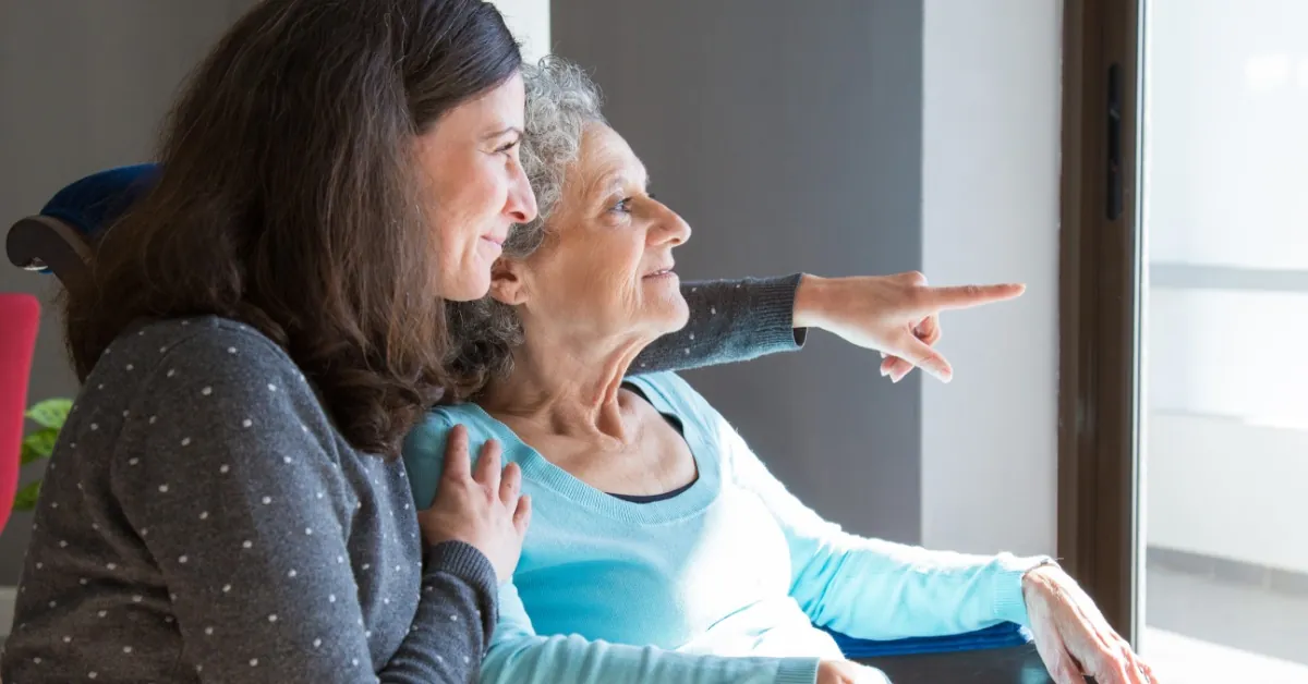 A middle-aged woman sits beside an older woman in a chair and points toward something outside a nearby window.